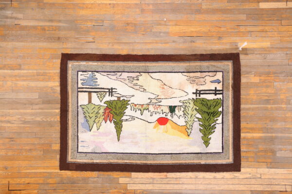 Multicolor hooked rug with a folk art landscape scene and thick brown border on hardwood floor planks.