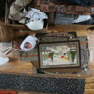 A man measures or examines several layered rugs on a wood floor next to large storage boxes.