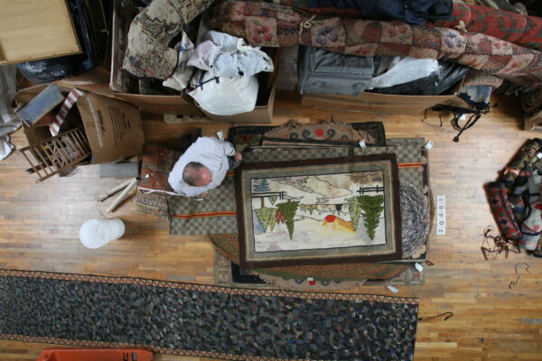 A man measures or examines several layered rugs on a wood floor next to large storage boxes.