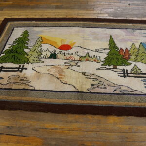 Hooked rug with a woodland scene and sunset pattern, framed by a dark border on worn wood planks.