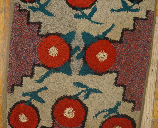 High-pile rug featuring red and cream flowers, teal shapes, and a speckled border resting on wood flooring.