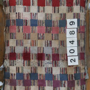 A textured patchwork rug with red, blue, pink, and beige checkered blocks is stacked over other rugs.