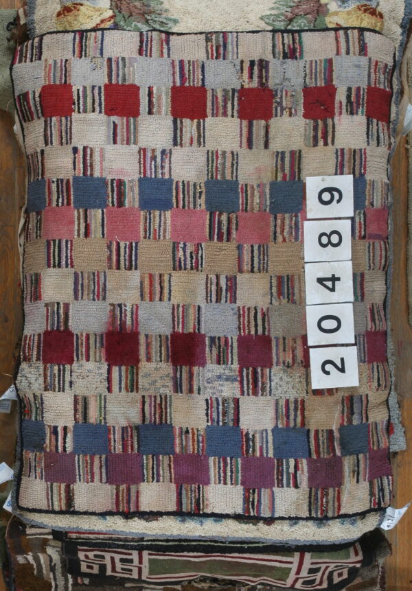 A textured patchwork rug with red, blue, pink, and beige checkered blocks is stacked over other rugs.