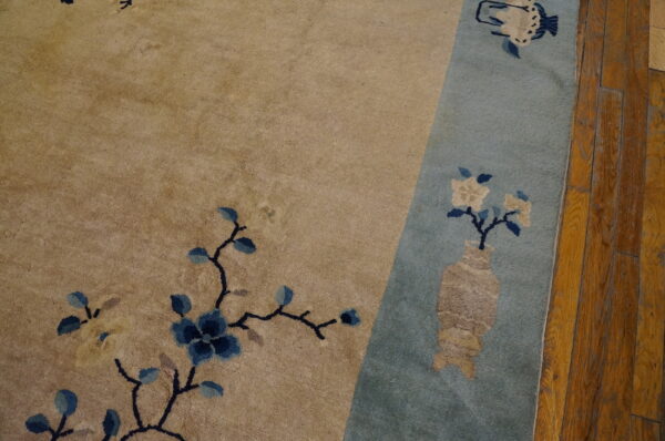 Beige rug field with blue floral vines borders a pale blue section containing a vase motif on wood planks.