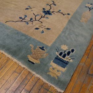 Tan rug with a teal border patterned with blue flowers and beige vessels resting on old hardwood flooring.