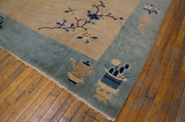 Tan rug with a teal border patterned with blue flowers and beige vessels resting on old hardwood flooring.