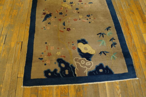 Tan pictorial rug with colorful birds and blossoms, bordered in deep blue, rests on golden wood planks.