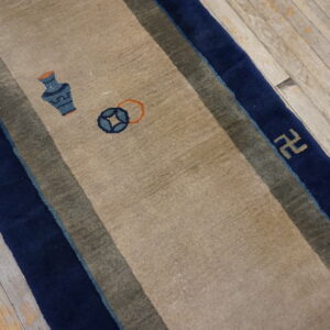 A beige runner rug with navy blue and olive borders displays vase and coin motifs on worn wooden flooring.
