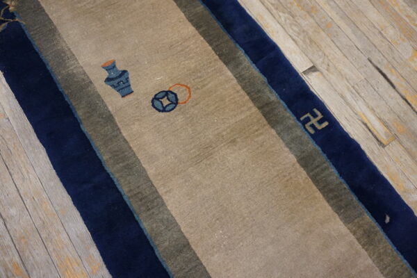 A beige runner rug with navy blue and olive borders displays vase and coin motifs on worn wooden flooring.