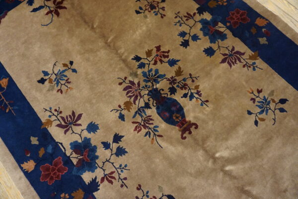 Tan rug detail showing a blue vase with colorful flowering branches and a wide indigo border.