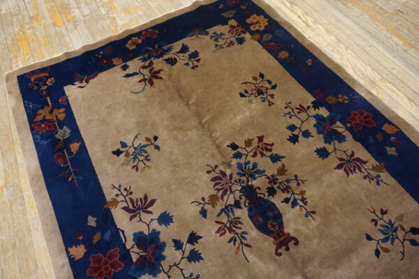Beige rug with deep blue border displays a central floral vase design on weathered wood planks.