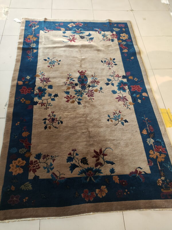 A beige and blue floral rug with a central vase motif sits on a white tiled floor.