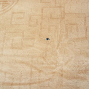 Tan plush rug detail with subtle carved geometric and scrolling patterns, plus a dark blue spot.