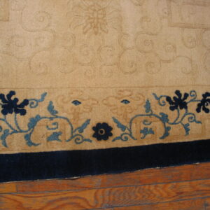 Tan rug detail showing embossed scrolling and a border with navy and teal floral patterns on wood flooring.