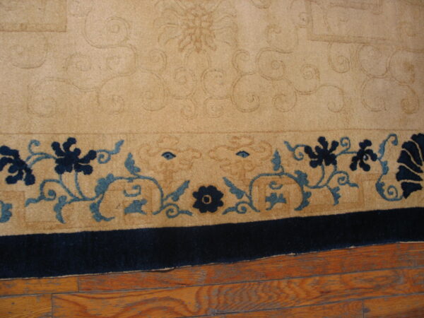Tan rug detail showing embossed scrolling and a border with navy and teal floral patterns on wood flooring.