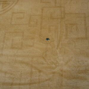 Tan plush rug with low-contrast geometric and scrolling patterns, showing a dark spot and part of a shoe.