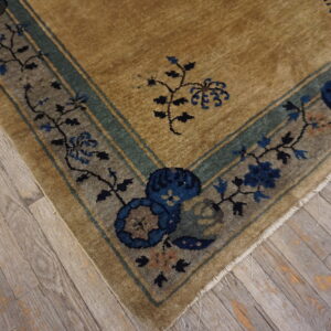 A low-pile rug corner featuring a tan field and indigo floral border resting on worn gray hardwood flooring.