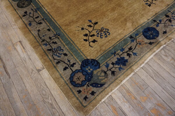 A low-pile rug corner featuring a tan field and indigo floral border resting on worn gray hardwood flooring.