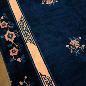 Deep blue high-pile rug featuring peach floral motifs and geometric borders rests on wood plank flooring.
