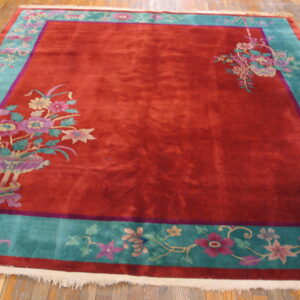 A red rug with a turquoise floral border and corner vase motifs rests on light wood floorboards.