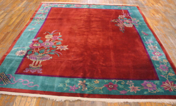 A red rug with a turquoise floral border and corner vase motifs rests on light wood floorboards.