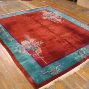Deep red plush rug featuring a wide teal border with pink floral motifs, set on worn wooden floorboards.