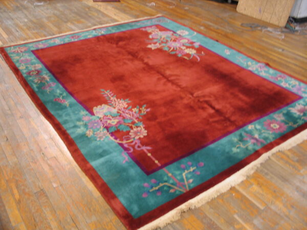 Deep red plush rug featuring a wide teal border with pink floral motifs, set on worn wooden floorboards.
