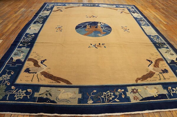 Large beige rug with a dark blue pictorial border featuring cranes and flowers on a polished wooden floor.