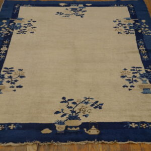 Beige field rug with a wide blue border and floral vase patterns resting on wood planks.