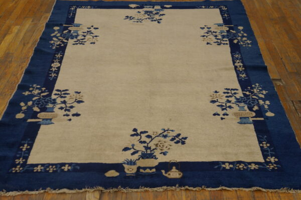 Beige field rug with a wide blue border and floral vase patterns resting on wood planks.