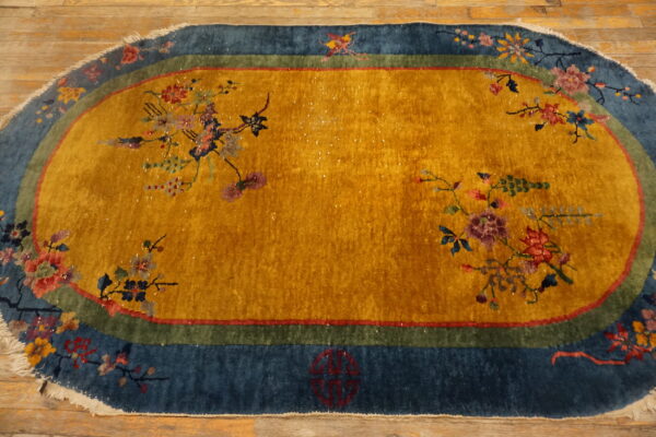 Gold oval rug with an ornate floral pattern and a wide blue and green border rests on wood planks.