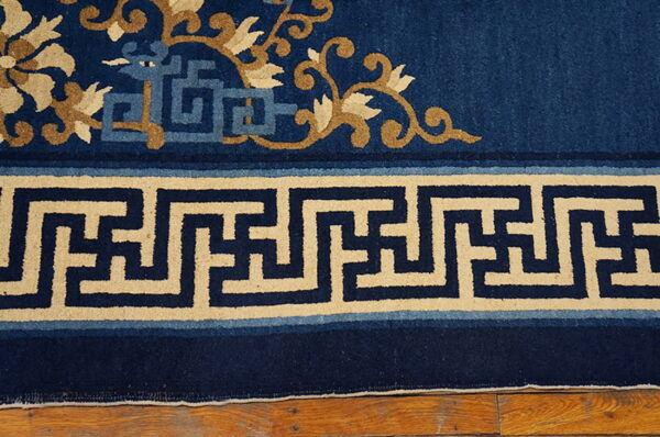 Low-pile navy blue rug with tan vines and a thick beige geometric key border resting on hardwood flooring.