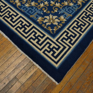 Navy blue rug corner with a beige Greek key border and floral designs rests on distressed wooden planks.