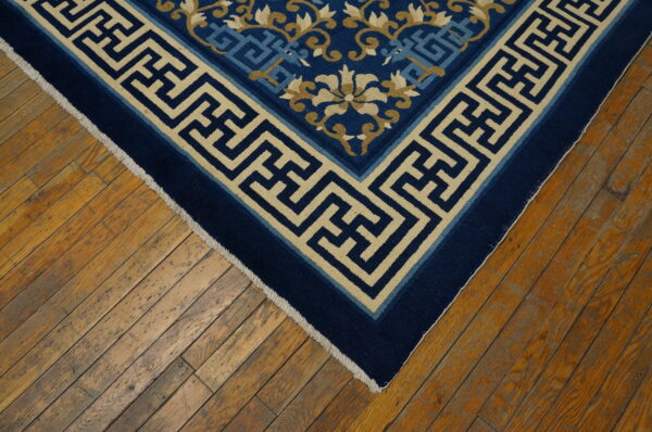 Navy blue rug corner with a beige greek key border and floral designs rests on distressed wooden planks.