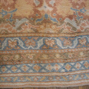 High-pile rug in muted peach, gold, and blue colors, showing bordered patterns over wood flooring.