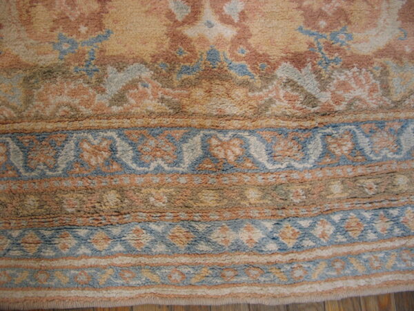 High-pile rug in muted peach, gold, and blue colors, showing bordered patterns over wood flooring.