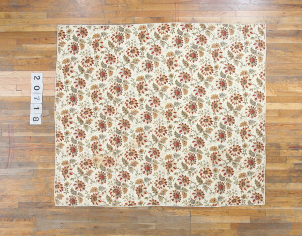 Cream rug with a repeating pattern of rust, gold, and green floral motifs laid on wide wooden floor planks.