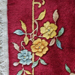 Deep red rug showing yellow and blue floral pile patterns next to a woven beige fringe.