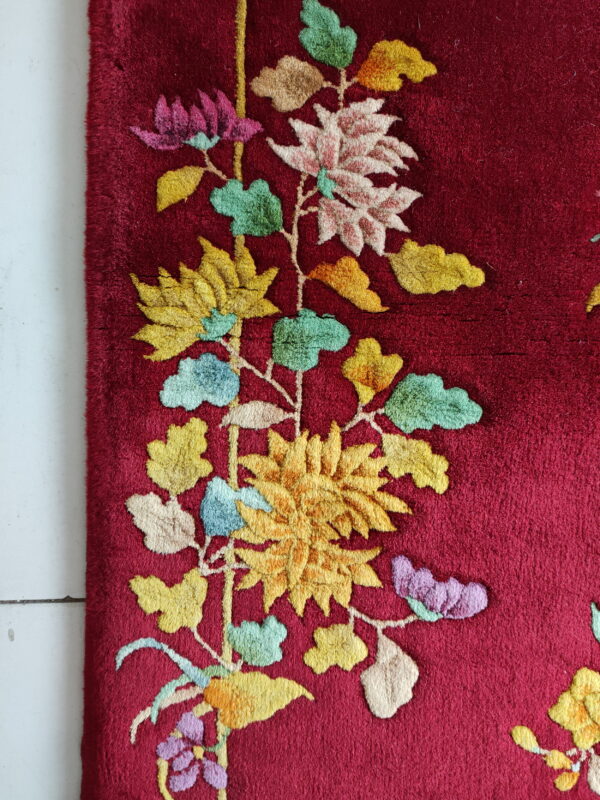A plush, deep red rug features a raised, vibrant floral and vine design on white flooring.