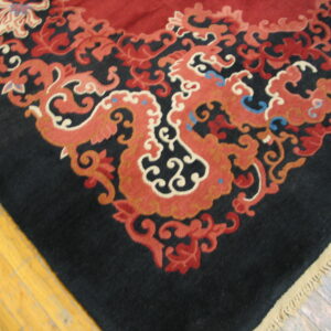 Black rug corner with intricate red, rust, and cream scrollwork borders a solid red field on wood floor.