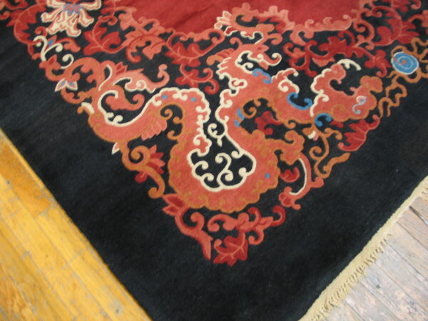 Black rug corner with intricate red, rust, and cream scrollwork borders a solid red field on wood floor.