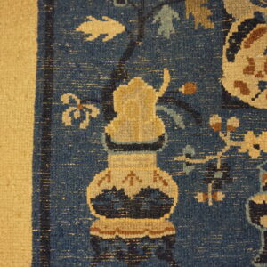 Blue patterned rug corner with a large stylized tan vase and wide solid tan border.