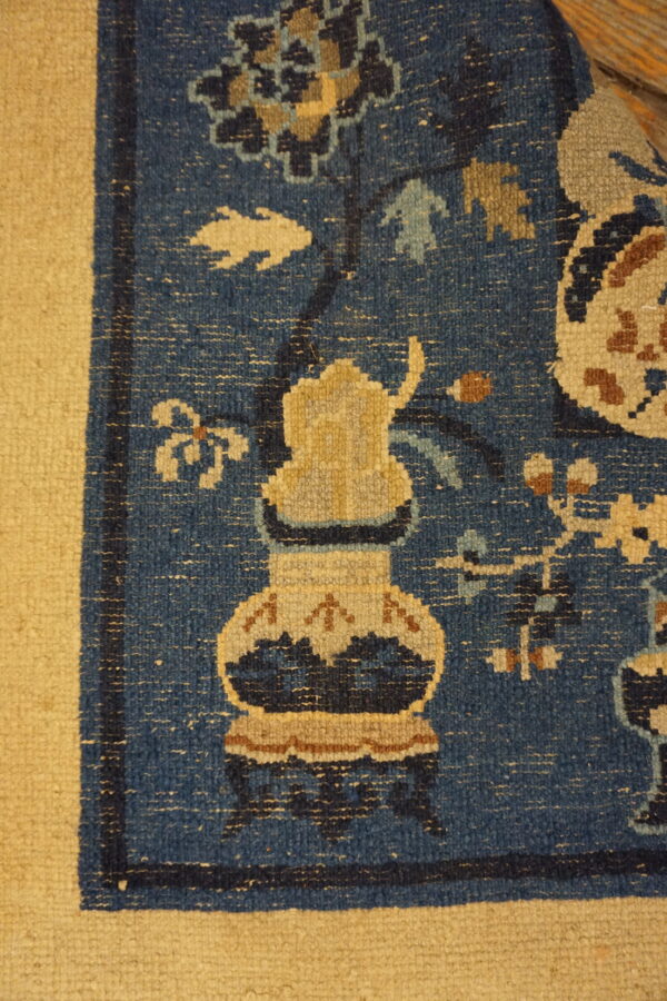 Blue patterned rug corner with a large stylized tan vase and wide solid tan border.