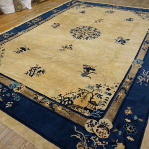 Large beige rug with deep blue borders and intricate botanical motifs lying on rough wooden planks.