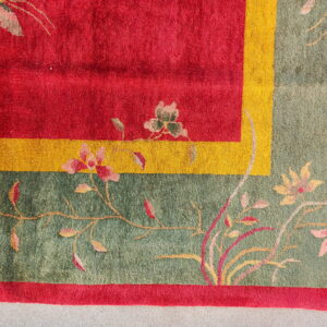 Red field rug corner with a yellow stripe and sage green floral border resting on gray flooring.