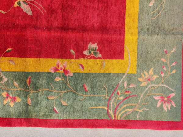 Red field rug corner with a yellow stripe and sage green floral border resting on gray flooring.