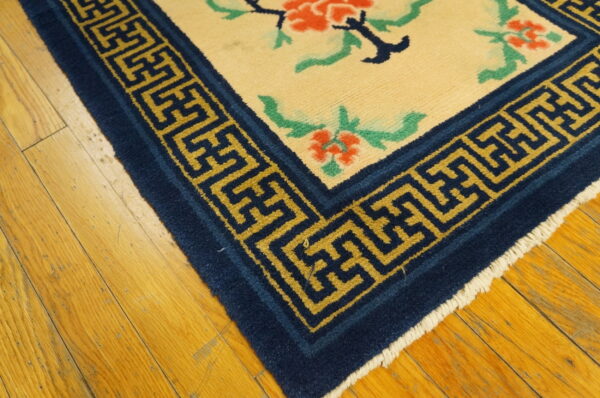 Cream area rug with orange and green florals and a dark blue geometric border rests on wood flooring.