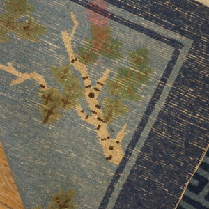 Folded blue flatweave rug shows a tree pattern and navy border resting on light wood flooring.