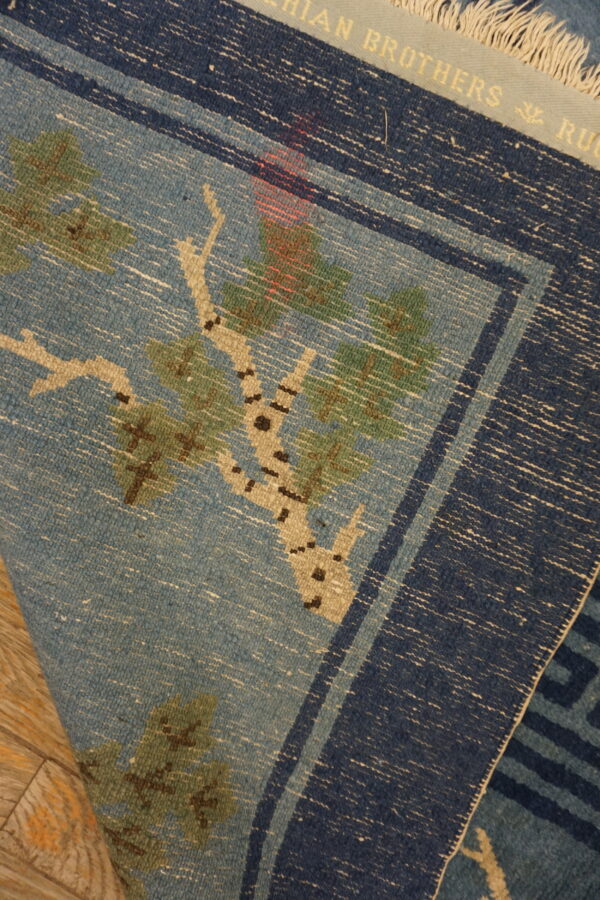 Folded blue flatweave rug shows a tree pattern and navy border resting on light wood flooring.