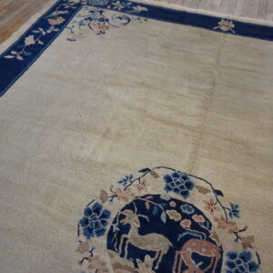 Light beige rug with a dark blue border and a medallion patterned with deer and flowers on wood flooring.